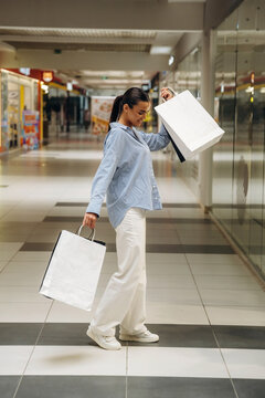 Female shopper in light blue shirt and white pants joyfully carries shopping bags while walking through a modern shopping mall with glass storefronts in the background