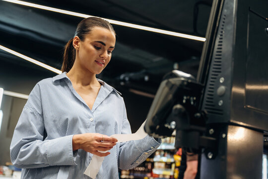 Young woman in a striped shirt holding a receipt while using a self-service kiosk in a modern grocery store with shelves of products visible in the background