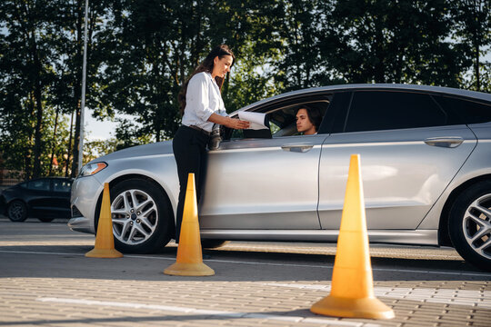 Female parking attendant in white shirt assists driver in gray sedan at outdoor parking lot with traffic cones and trees in the background