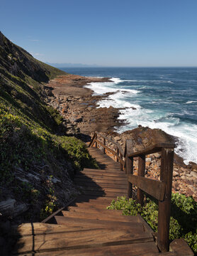Scenic view from the Point Hike trail in Robberg Nature Reserve, South Africa, featuring a wooden boardwalk with handrails leading down a rugged coastline towards a wild ocean and distant bay.