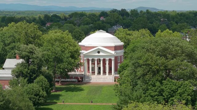 Charlottesville, Virginia, aerial orbiting drone video rising high above lush green trees to reveal the University of Virginia Rotunda and campus on a sunny summer day.