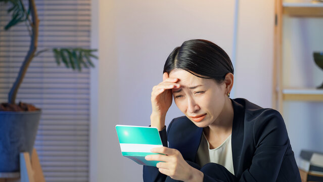 暗い部屋で通帳を見て落ち込むミドル女性。貯金、不安、心配、銀行、家計、投資、資産運用、物価、増税