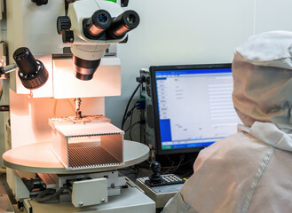 A technician in a cleanroom uses a binocular microscope to inspect small electronic components, with a monitor displaying process data nearby. © tai