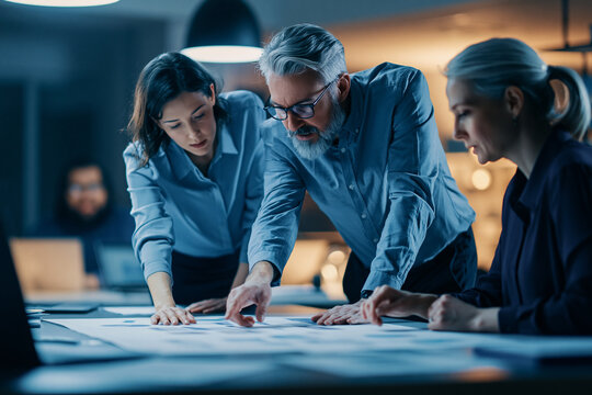 Three professional colleagues collaborate in a dimly lit office leaning over a large document on a table to analyze complex data and strategize together