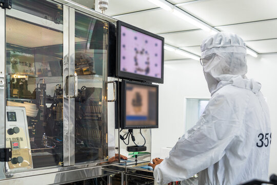 A worker in a cleanroom operates precision manufacturing equipment, monitoring work on two display screens.