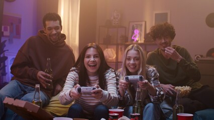 Young people sitting on couch and playing video games. Friends having fun and holding snacks. Company of people smiling and gesturing. The girl gives high five to boy. Evening leisure of youth. © VAKSMANV