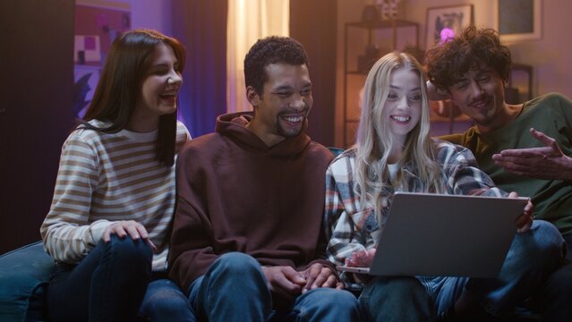Blonde girl is holding laptop. Company of young people is sitting on couch and laughing. Friends look at screen device and communicate. Evening entertainment for young people.