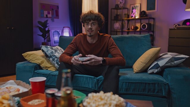Front view shoot. Curly head guy is sitting on coach. Young man is holding joystick and playing video games. Young adult having leisure at background of modern room and snacks.
