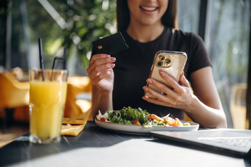 Obraz premium Young woman using smartphone and credit card to pay for healthy meal at a restaurant table with salad and orange juice in a bright, modern setting