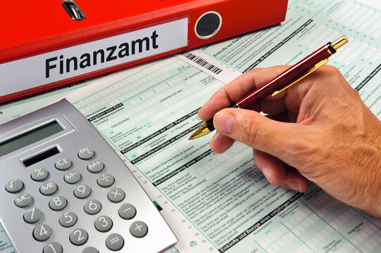 Eine Hand mit Kugelschreiber &uuml;ber einem Formular f&uuml;r das Finanzamt, dahinter liegt eien Aktenordner mit Finanzamt beschriftet.