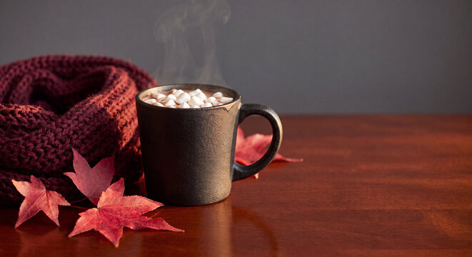 Cozy autumn scene with steaming hot cocoa, knitted scarf and maple leaves on wooden table