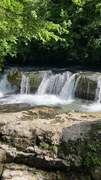 clear river water pours over a low rocky waterfall beneath lush green forest foliage, creating a refreshing natural stream scene with splashing water in peaceful summer woodland