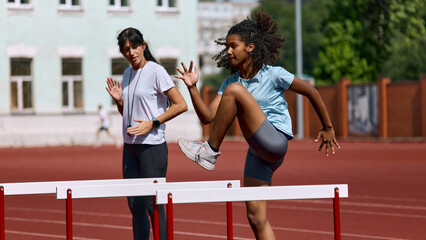 Fototapeta premium Teenage female athlete practicing hurdle approach while adult female coach instructs. Concept of hurdle preparation routine, youth athlete instruction, summer sport skill building.