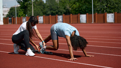 Fototapeta premium Teenage female runner in crouched start position with adult female coach assisting. Concept of starting position setup, sprint technique preparation, summer sport coaching.