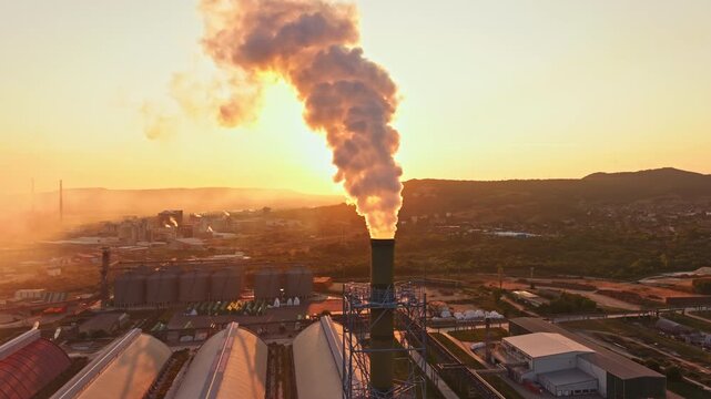 A large plume of smoke is seen coming from a factory chimney as the sun sets in the background. Industrial buildings are visible in the foreground and hills are far away.