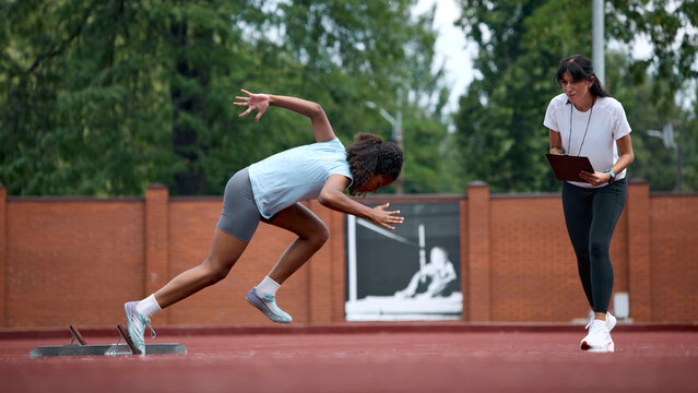 Teenage female runner launching from starting blocks while adult female coach monitors performance. Concept of starting block technique, stopwatch performance analysis, summer sport instruction.
