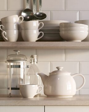 White ceramic teapot, cups, bowls and plates arranged on bright kitchen shelves