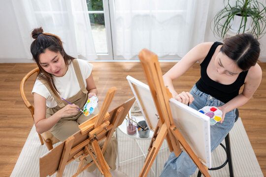 Two young Asian women painting together in a bright home studio during the weekend