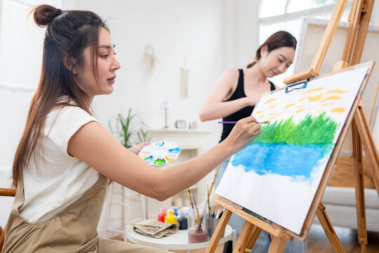 Two young Asian women painting a colorful landscape on canvas together