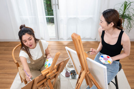 Two young Asian women painting together in a bright home studio during the weekend