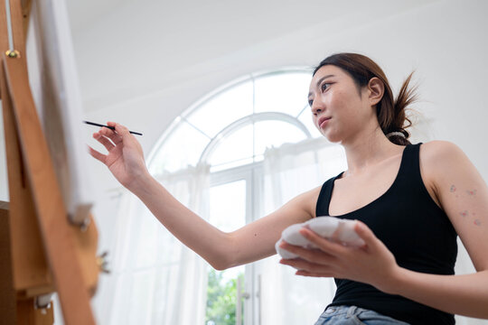 Young Asian woman painting on canvas in a bright home art studio during the weekend