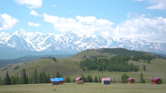 Breathtaking establishing shot of a remote countryside holiday resort featuring simple wooden huts on a green meadow against a magnificent panoramic view of epic snowy mountain peaks.