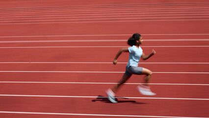 Fototapeta premium Side view of Black teenage female runner accelerating across red running track with motion blur. Concept of sprint speed development, youth athletics training, summer sport performance.