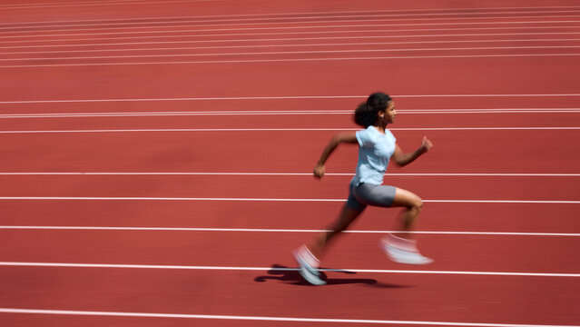 Side view of Black teenage female runner accelerating across red running track with motion blur. Concept of sprint speed development, youth athletics training, summer sport performance.