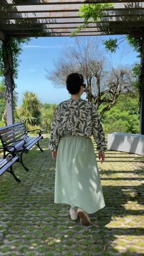woman in a patterned blouse and flowing skirt walks away beneath a leafy garden pergola, capturing a calm summer moment of leisure, solitude and slow outdoor living