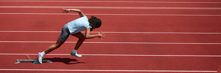 Fototapeta premium Teenage female runner launching from low crouched sprint start on track. Concept of sprint start technique, athletic readiness, summer sport performance. Banner