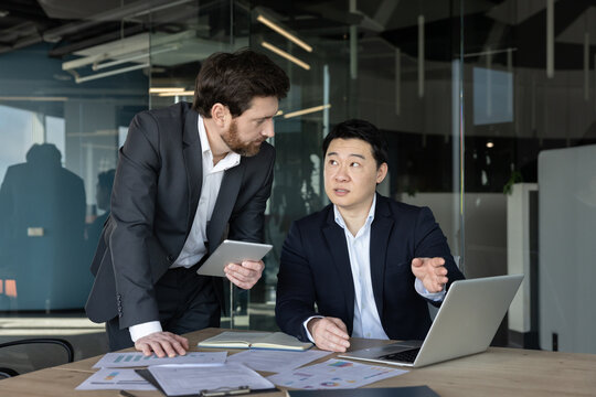 Business professionals collaborating in a meeting, reviewing financial charts and reports on a laptop and tablet, actively discussing strategy and project planning in a modern office environment