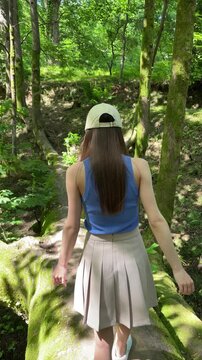 young woman in a cap and pleated skirt walks along a shaded woodland trail, exploring a green forest during a quiet summer hike surrounded by mossy trees and ferns