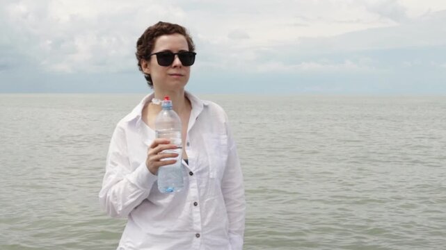 woman recovering from cancer holds drinking water by the ocean on a summer break, highlighting hydration, sun protection, life after treatment and resilient travel