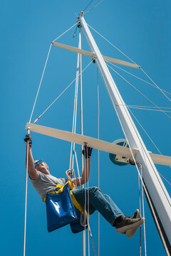 Caucasian Male working up the mast of a sailing yacht, with rope and bosun's chair on a sunny day with blue sky 