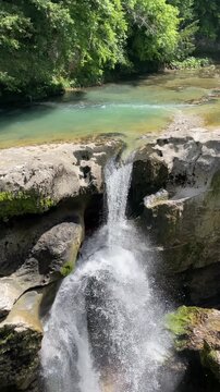 clear river water drops through a narrow opening in pale limestone rocks, creating a powerful waterfall with bright spray, mossy textures and a hidden forest gorge