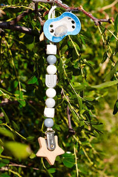 Lost pacifier hanging from a tree branch, attached to a colorful beaded strap with a wooden star clip, surrounded by green leaves in a natural outdoor setting