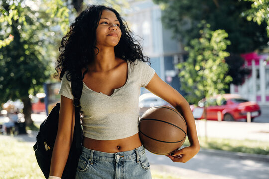 Young African American woman with curly hair wearing a gray t-shirt and denim shorts holds a basketball while walking in a sunny urban park setting