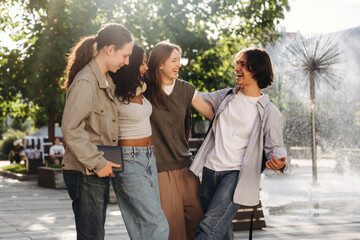 Fototapeta premium Group of four young adults laughing and enjoying time together outdoors near a fountain in a park with trees and sunlight filtering through the leaves