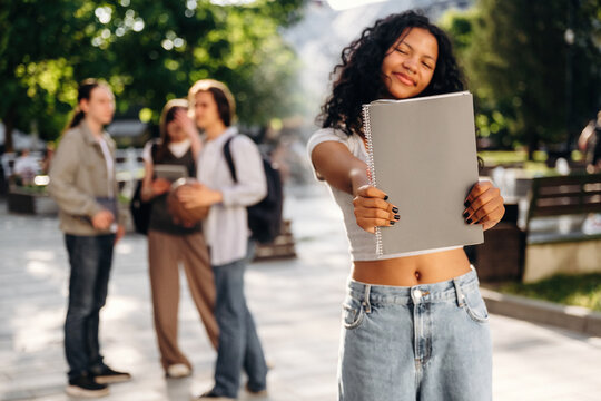 Young woman with curly hair holds a notebook in a park, smiling at the camera while friends chat in the background under bright sunlight and green trees