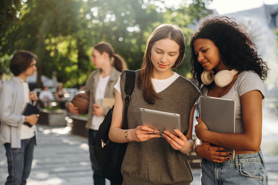 Two young women, one with brown hair and the other with curly black hair, examine a tablet together in a sunny outdoor park setting with students in the background