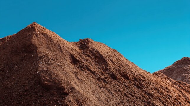 Large pile of brown construction sand and dirt against clear blue sky. Earth mound for building works and development concept. Raw material for civil engineering and infrastructure.