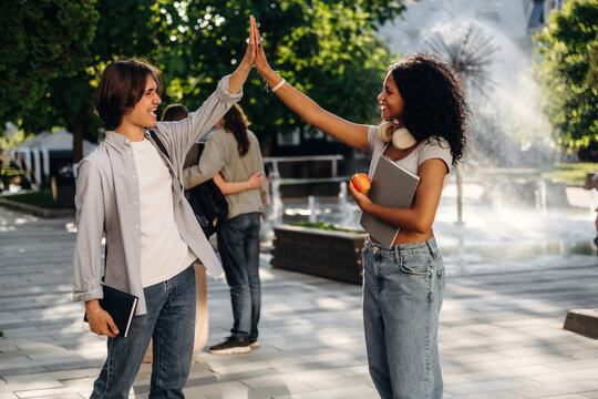 Young Caucasian male and African American female students high-five each other in a park setting with a fountain and greenery in the background