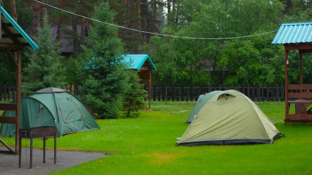 Slow camera push in revealing a scenic forest campsite with camping tents and wooden shelters on a green lawn surrounded by tall pine trees in the morning.