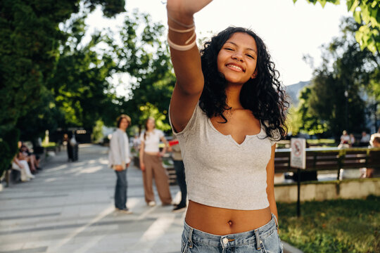 Young woman with curly hair wearing a gray crop top and denim shorts smiles and waves in a park with people and greenery in the background