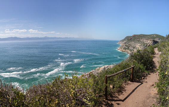 High resolution stitched panorama of Robberg Nature Reserve, South Africa, featuring a wide angle panoramic view of a coastal hiking trail with wooden handrails along steep cliffs and wild ocean.
