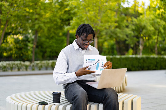 A smiling african american man wearing glasses and business casual attire reviews a financial report with charts and graphs while working on his laptop outside on a bench