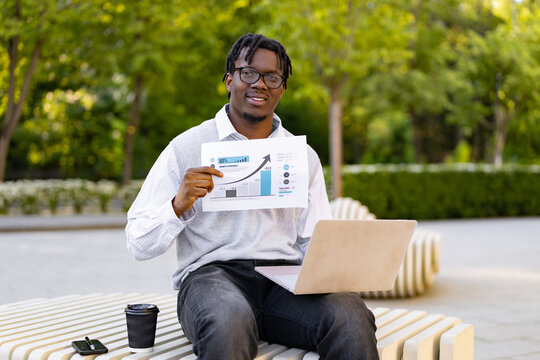 A smiling african american man in glasses sits outdoors with a laptop and a coffee cup, presenting a document showing a positive business growth chart