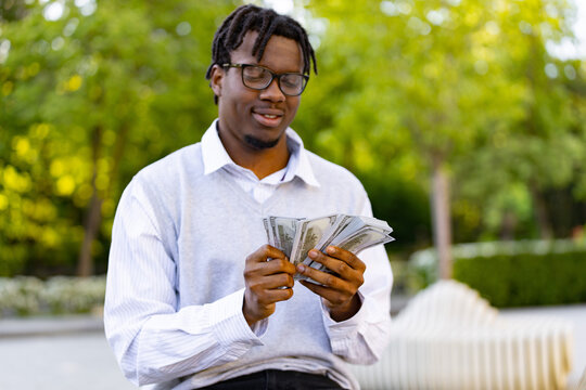 A smiling african american man wearing glasses holds and counts a fan of US dollar bills while sitting outside in a park setting