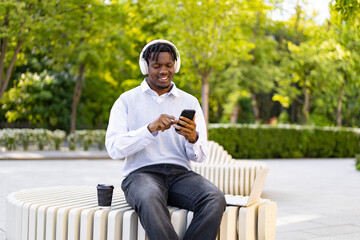 Obraz premium A smiling young African American man wearing headphones sits on a modern bench in a park, looking at his phone with a laptop nearby