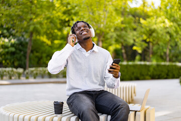 Obraz premium A young African American man wearing headphones and holding a smartphone enjoys music while sitting on a bench in a park with a laptop and coffee nearby